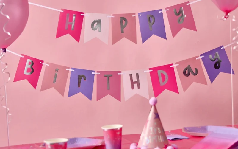 A pink birthday party table decorated with a pink, lilac and silver ‘Happy Birthday’ bunting banner hanging against a pale pink background, with matching party cups, plates, balloons and a party hat arranged below.