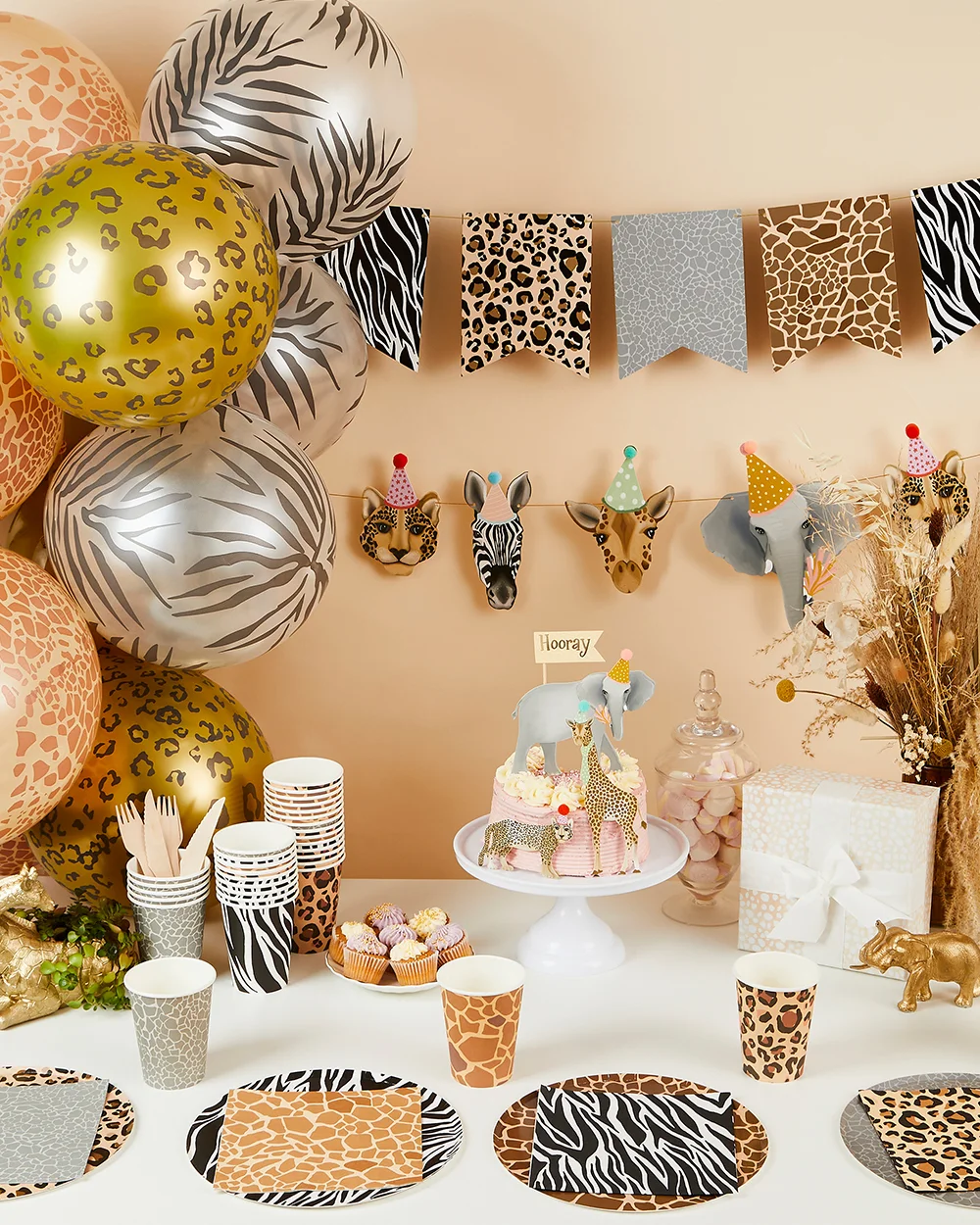 A safari-themed party table decorated with animal-print partyware, including zebra, leopard, giraffe and textured-pattern plates, cups and napkins. The display features a jungle-inspired cake topped with elephant and giraffe decorations, cupcake treats, metallic animal-print balloons, animal face bunting, and safari-themed table accessories arranged against a neutral beige backdrop.