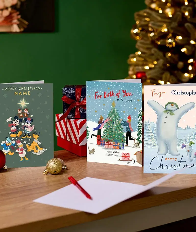 Selection of Christmas cards, including Disney and personalised designs, displayed on a wooden table with wrapped presents and a decorated Christmas tree in the background. Selection of Christmas cards, including Disney and personalised designs, displayed on a wooden table with wrapped presents and a decorated Christmas tree in the background.