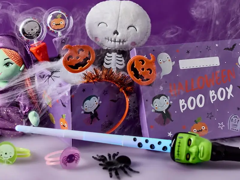 Halloween party accessories including a Boo Box, skeleton decoration, pumpkin headband, glow wand, novelty sweets and spooky themed toys on a purple background.