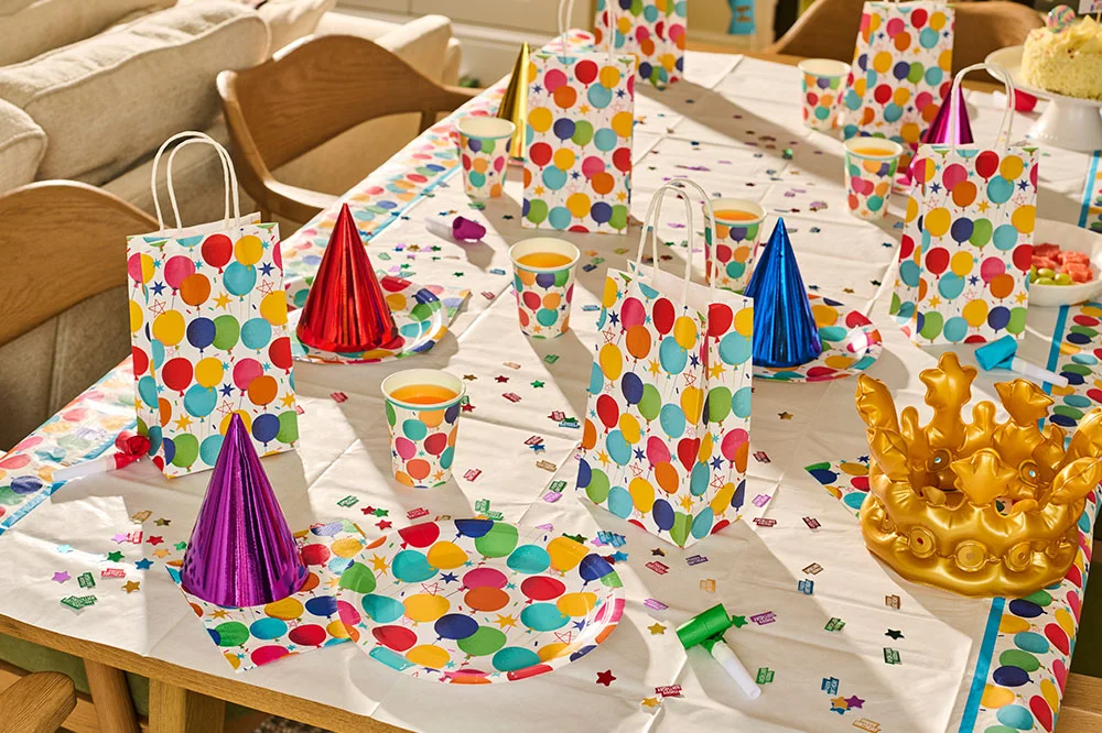 A children's birthday party table decorated with balloon-print partyware, including matching gift bags, paper cups, plates and napkins. Colourful confetti, metallic party hats, party blowers and a gold crown centrepiece are arranged across the table, creating a vibrant birthday celebration setup.