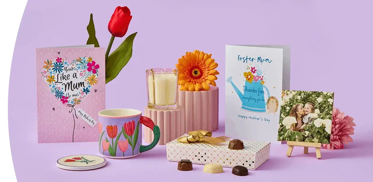 A cheerful Mother’s Day display featuring two greeting cards, a red tulip, a floral mug with a matching coaster, a candle, a gift box with assorted chocolates, and a small photo plaque arranged on a lilac background. A cheerful Mother’s Day display featuring two greeting cards, a red tulip, a floral mug with a matching coaster, a candle, a gift box with assorted chocolates, and a small photo plaque arranged on a lilac background.