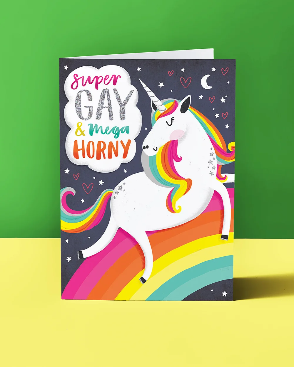 A Pride greeting card featuring a white unicorn with a rainbow mane standing over a rainbow, surrounded by stars and hearts, with bold multicoloured text reading ‘Super Gay & Mega Horny,’ displayed against a green and yellow background.