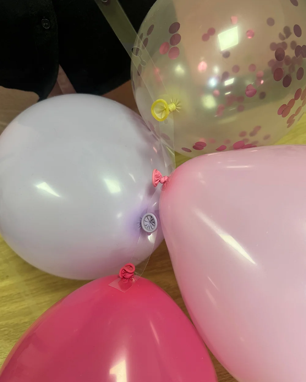 A close-up of four inflated balloons tied together to form a balloon cluster, including pink, lilac, white and clear balloons filled with pink confetti. The balloons are joined at their tied necks, demonstrating how balloon groups are assembled for use in a balloon arch decoration. A close-up of four inflated balloons tied together to form a balloon cluster, including pink, lilac, white and clear balloons filled with pink confetti. The balloons are joined at their tied necks, demonstrating how balloon groups are assembled for use in a balloon arch decoration.