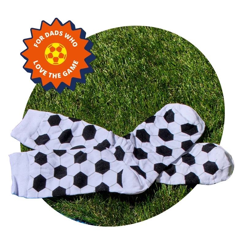 A pair of novelty socks with a black and white football pattern, laid out on a circular patch of green grass, displayed against an orange background with a ‘For dads who love the game’ badge. A pair of novelty socks with a black and white football pattern, laid out on a circular patch of green grass, displayed against an orange background with a ‘For dads who love the game’ badge.