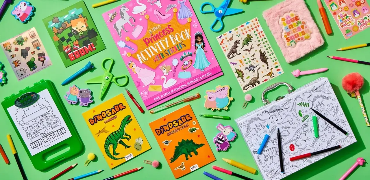 A colorful assortment of children's activity books and stationery items are arranged on a green background. The items include a pink 'Princess Activity Book with Stickers', two dinosaur-themed activity books, a Minecraft-themed book, various stickers, colored pencils, scissors, and other craft supplies.