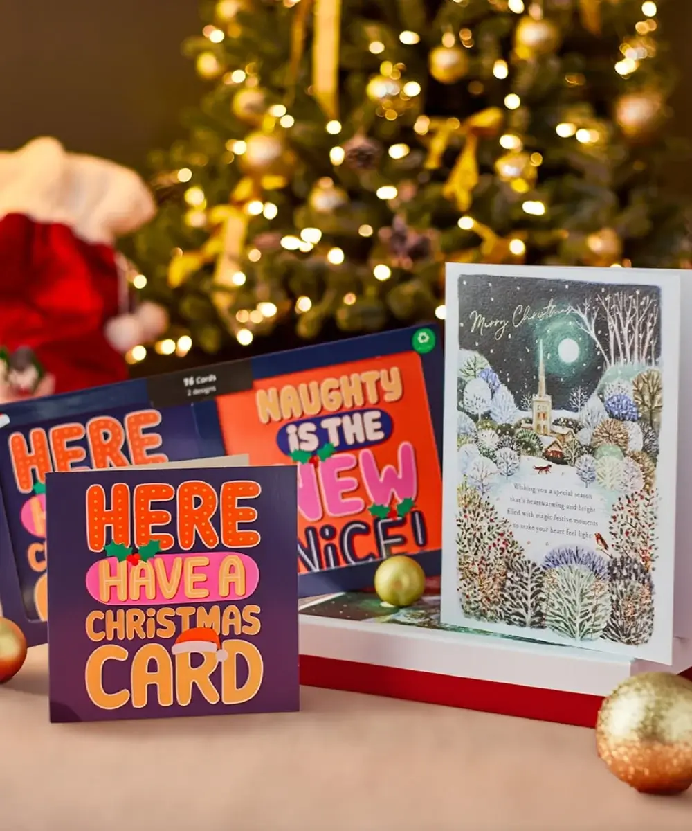 Festive scene with a decorated Christmas tree and three Christmas cards featuring bold text, playful messages, and a snowy village illustration. Festive scene with a decorated Christmas tree and three Christmas cards featuring bold text, playful messages, and a snowy village illustration.