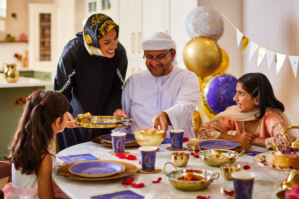 AA family seated at a dining table sharing food during a festive celebration, with decorative plates, bowls, rose petals, and balloons arranged around the table.