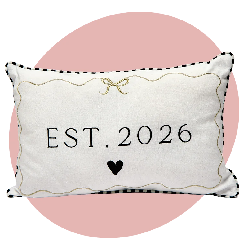 A rectangular cream cushion with black trim and gold decorative stitching, featuring the text ‘EST. 2026’ and a small black heart in the centre. A rectangular cream cushion with black trim and gold decorative stitching, featuring the text ‘EST. 2026’ and a small black heart in the centre.