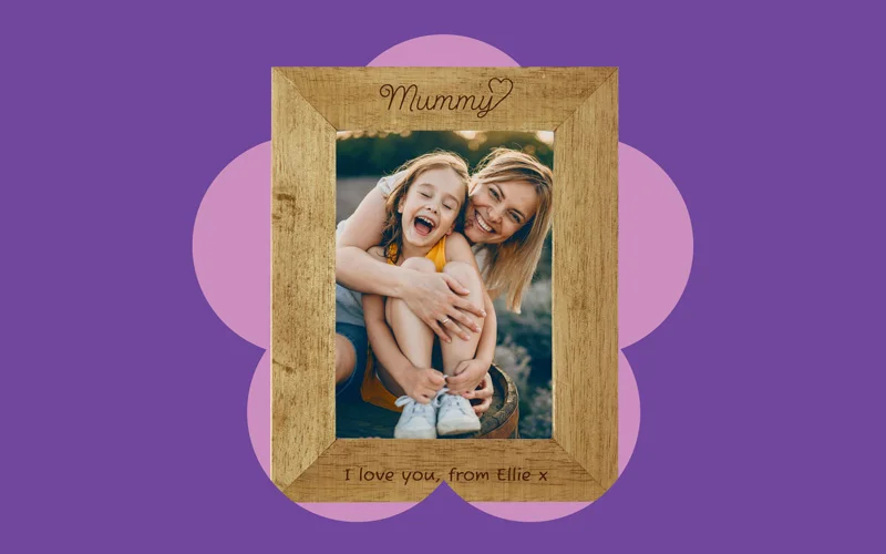 A wooden photo frame engraved with the word ‘Mummy’ at the top and a personalised message at the bottom, displayed against a purple background with a pink flower shape.
