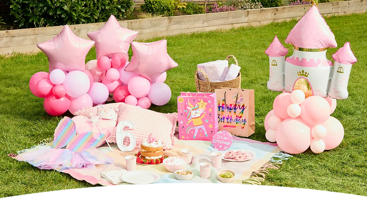 A princess-themed birthday picnic set up on a garden lawn, featuring a pastel pink picnic blanket with party tableware, cupcakes, snacks and drinks. The display includes pink balloon clusters with star-shaped foil balloons, a pink castle-shaped balloon decoration, princess dress-up accessories, birthday gift bags, wrapped presents, and white number 6 decorations arranged around the picnic area. A princess-themed birthday picnic set up on a garden lawn, featuring a pastel pink picnic blanket with party tableware, cupcakes, snacks and drinks. The display includes pink balloon clusters with star-shaped foil balloons, a pink castle-shaped balloon decoration, princess dress-up accessories, birthday gift bags, wrapped presents, and white number 6 decorations arranged around the picnic area.