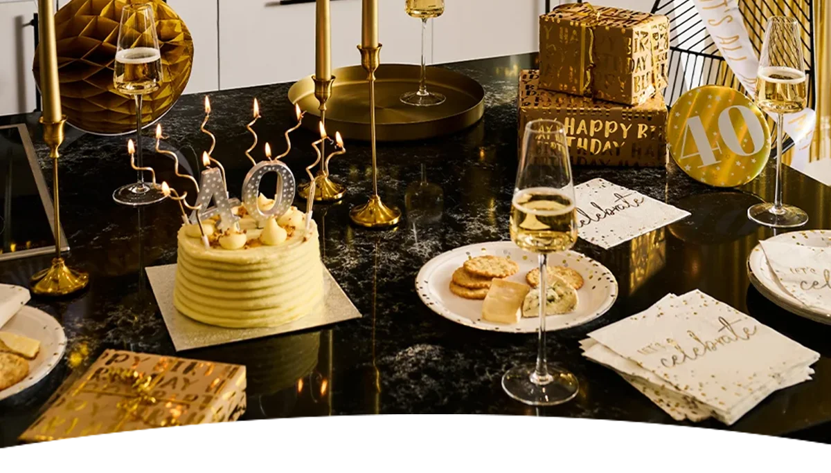 A black, gold and white milestone birthday table setting featuring a celebration cake topped with lit number 40 candles and gold spiral candles. The table is decorated with gold candlesticks, champagne flutes, wrapped gifts, black-and-gold partyware, and a gold ‘40’ balloon, creating an elegant 40th birthday celebration display. A black, gold and white milestone birthday table setting featuring a celebration cake topped with lit number 40 candles and gold spiral candles. The table is decorated with gold candlesticks, champagne flutes, wrapped gifts, black-and-gold partyware, and a gold ‘40’ balloon, creating an elegant 40th birthday celebration display.