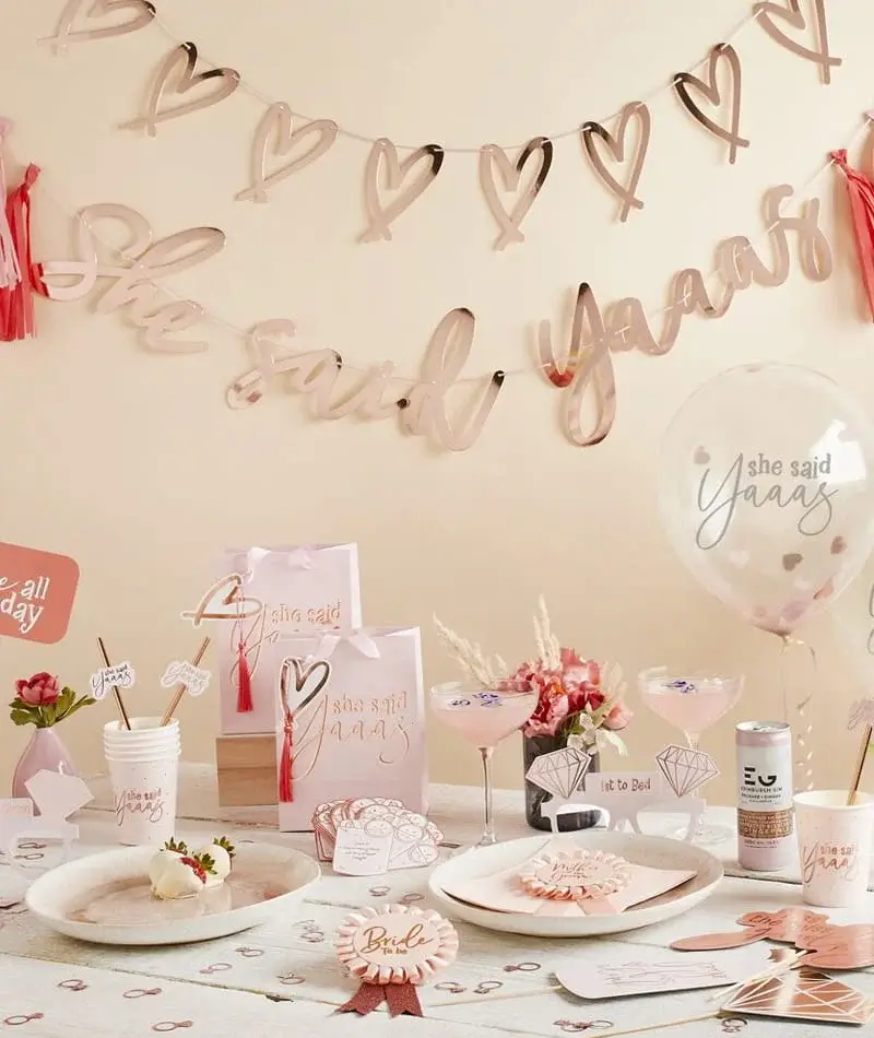 Hen party table setup with she said yaaass banner, themed decorations, drinks, plates, and party accessories in blush pink tones