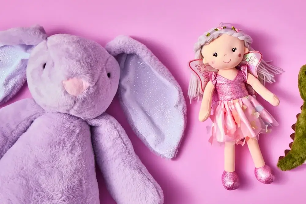 Plush purple bunny and fairy doll in a pink dress with wings on a solid pink background.