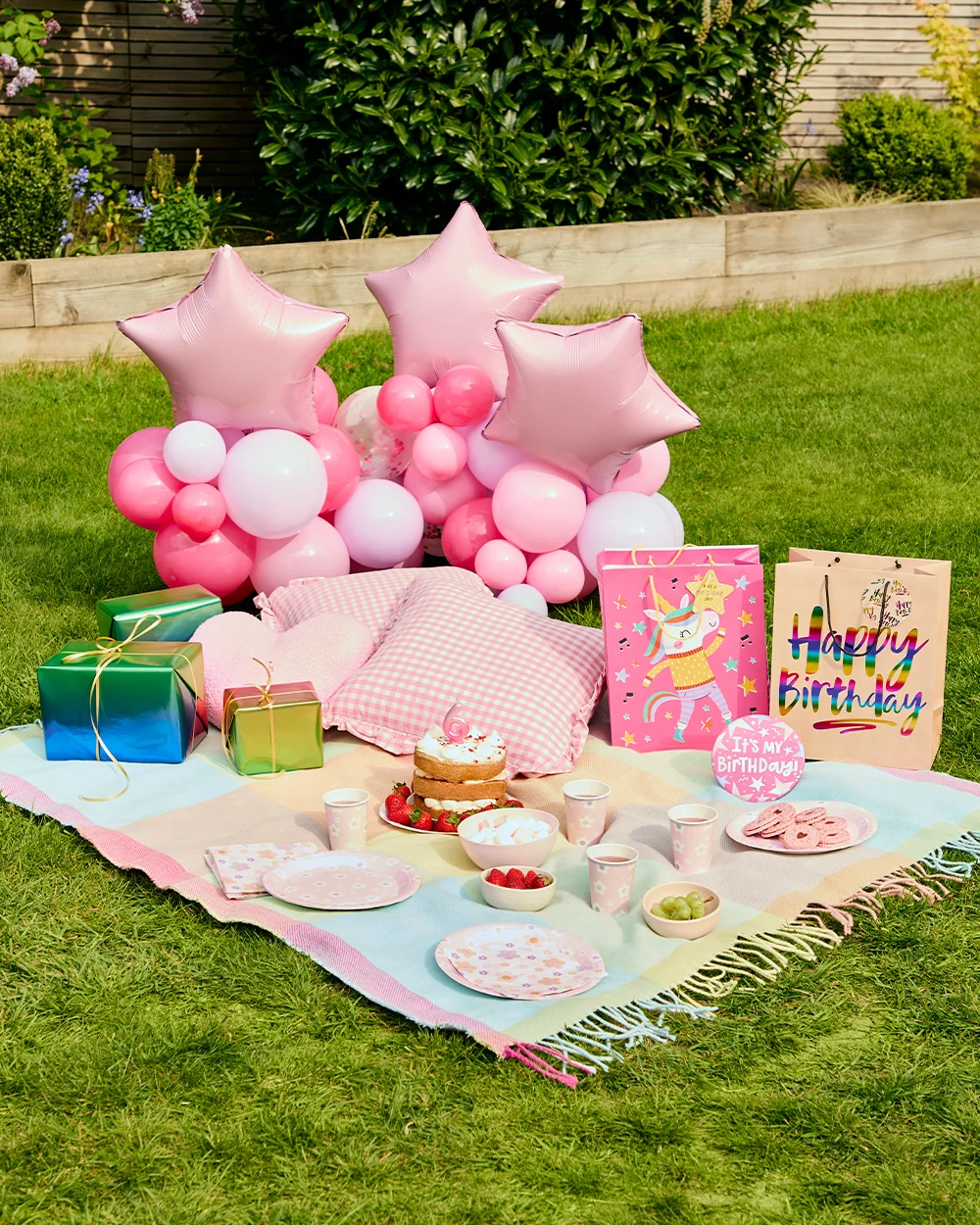 A pastel pink birthday picnic setup arranged on a lawn, featuring a picnic blanket laid with floral party plates, cups, snacks and a small celebration cake topped with a number 6 candle. The display includes pink star-shaped foil balloons, a pink balloon cluster backdrop, gingham cushions, wrapped gifts, and colourful birthday gift bags decorated with unicorn and ‘Happy Birthday’ designs, creating a whimsical outdoor party scene.
