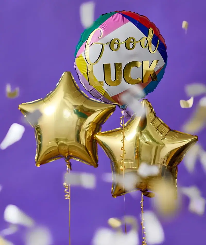 Good luck balloon bouquet featuring a colourful round balloon and gold star-shaped balloons, surrounded by falling confetti against a purple background.