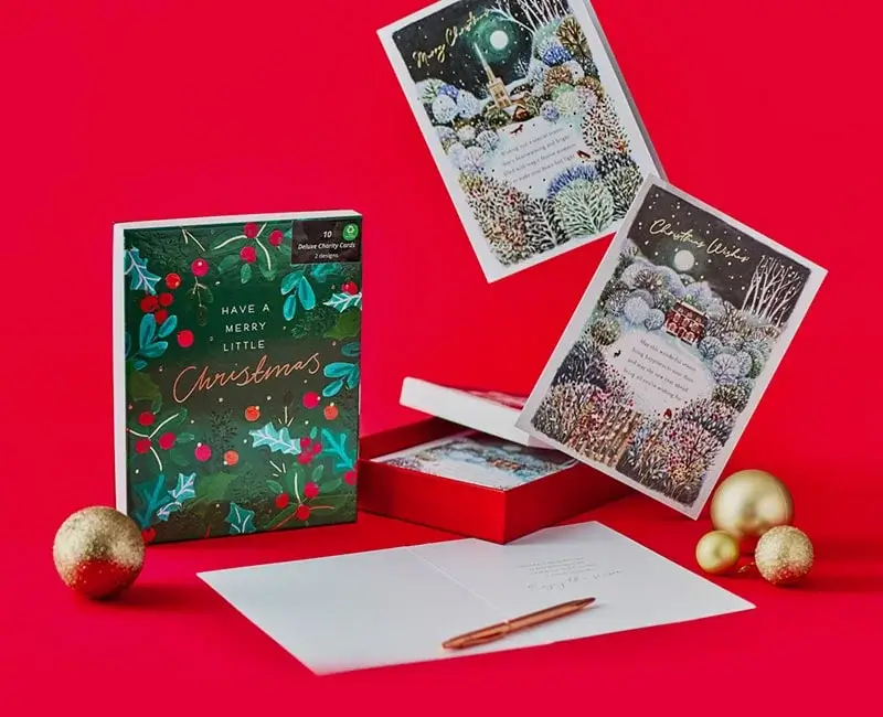 Set of Christmas cards on a bright red background, including a box labeled Have a Merry Little Christmas with festive foliage and berries, two propped-up cards showing snowy winter scenes with a church and a house, gold ornaments, and an open card with an envelope and pen.
