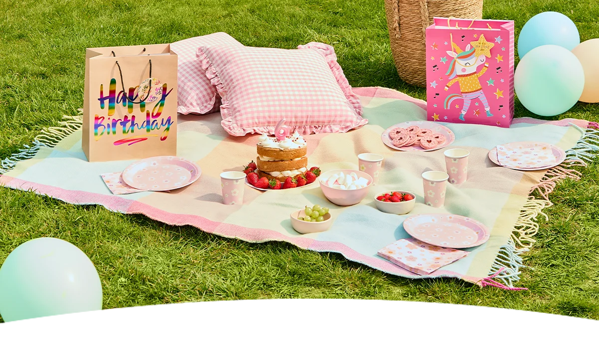 A pastel-themed children's garden party picnic set up on a lawn, featuring a pink-and-white blanket with gingham cushions, pink floral party plates, cups and napkins, and a small birthday cake topped with a number 6 candle. The display includes bowls of strawberries, grapes and marshmallows, pastel balloons, and colourful gift bags decorated with ‘Happy Birthday’ lettering and a unicorn design, creating a whimsical outdoor birthday celebration scene. Add alt text here