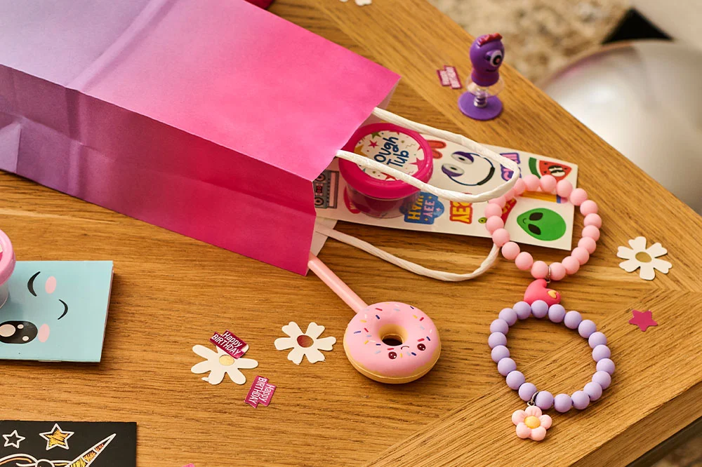 A close-up of a pink party favour bag spilling out children's party bag fillers, including a tub of dough, a colourful sticker sheet, a novelty spring toy, a donut-shaped toy and pastel beaded bracelets. Birthday-themed confetti and daisy-shaped decorations are scattered across the wooden tabletop. A close-up of a pink party favour bag spilling out children's party bag fillers, including a tub of dough, a colourful sticker sheet, a novelty spring toy, a donut-shaped toy and pastel beaded bracelets. Birthday-themed confetti and daisy-shaped decorations are scattered across the wooden tabletop.