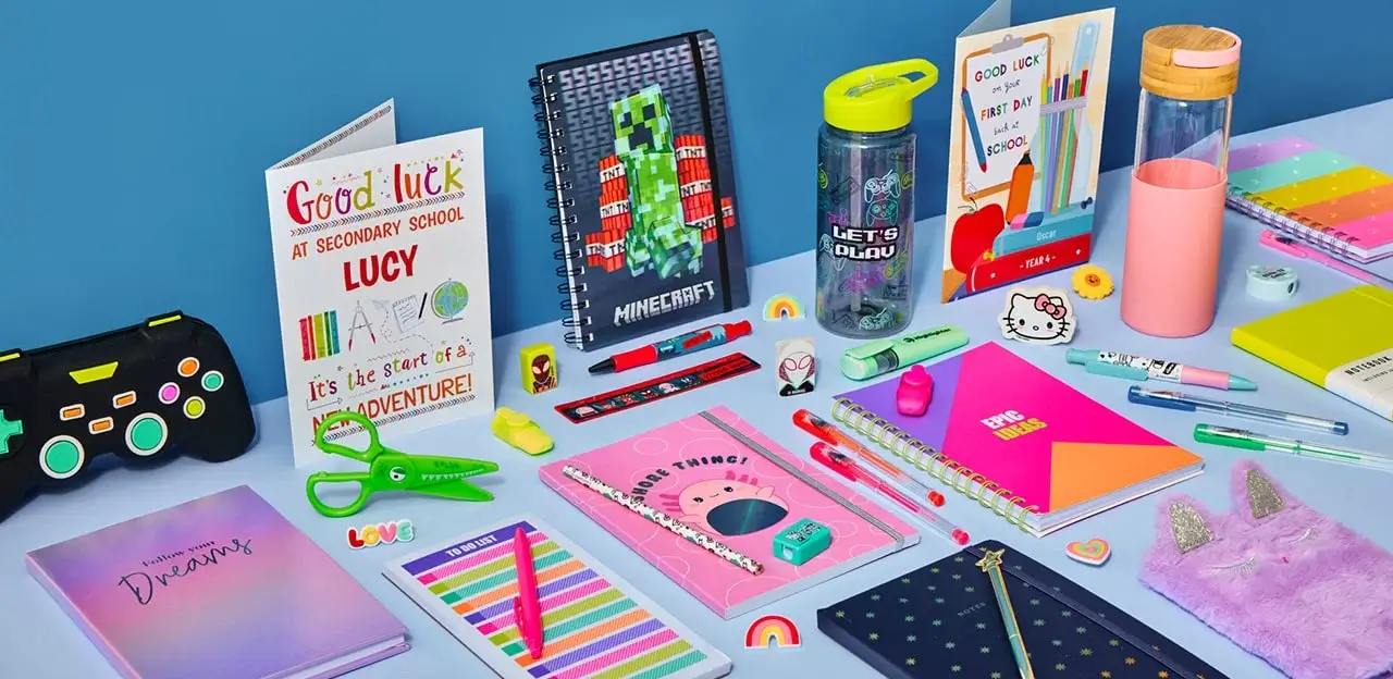 A colorful assortment of school supplies and stationery on a light blue surface, including notebooks, greeting cards, pens, pencils, a game controller, and a green water bottle.
