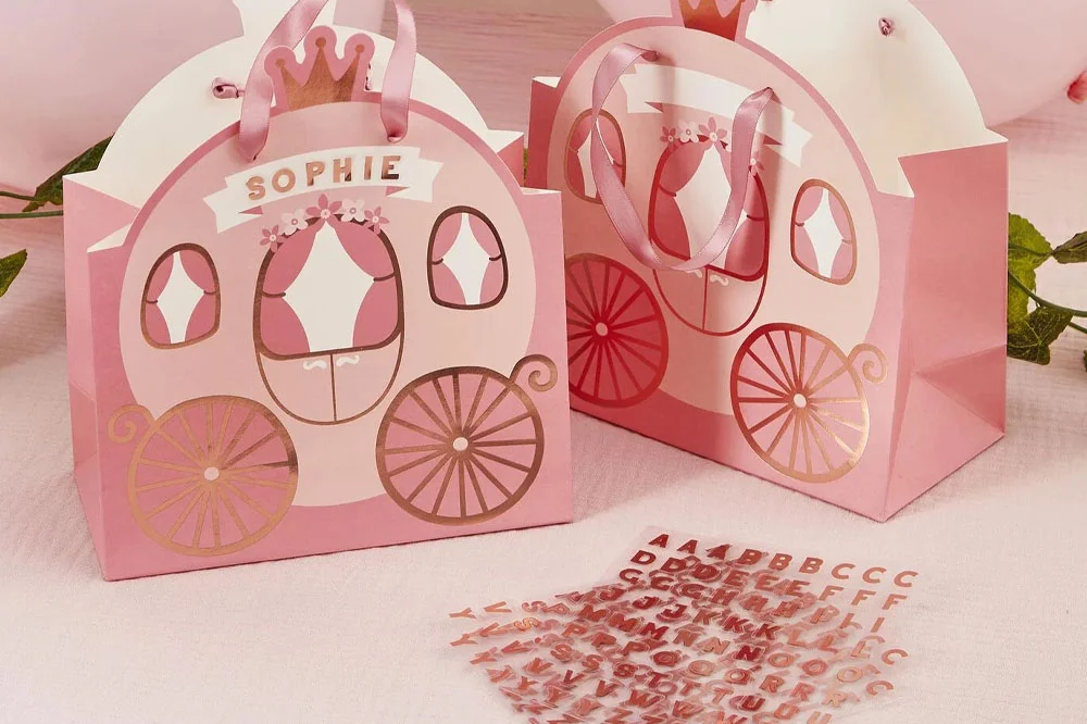 Princess-themed party favour boxes designed as pink fairytale carriages with gold foil details and ribbon handles, displayed alongside a sheet of pink alphabet stickers for personalising names and messages. Princess-themed party favour boxes designed as pink fairytale carriages with gold foil details and ribbon handles, displayed alongside a sheet of pink alphabet stickers for personalising names and messages.