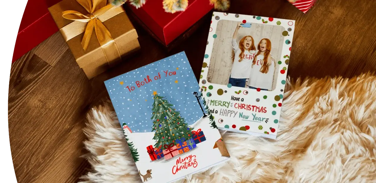 Two Christmas cards placed on a fluffy rug next to wrapped gifts. One card features a decorated Christmas tree with presents and the text To Both of You Merry Christmas, while the other has a festive design with colorful dots and the message Have a Merry Christmas and a Happy New Year.