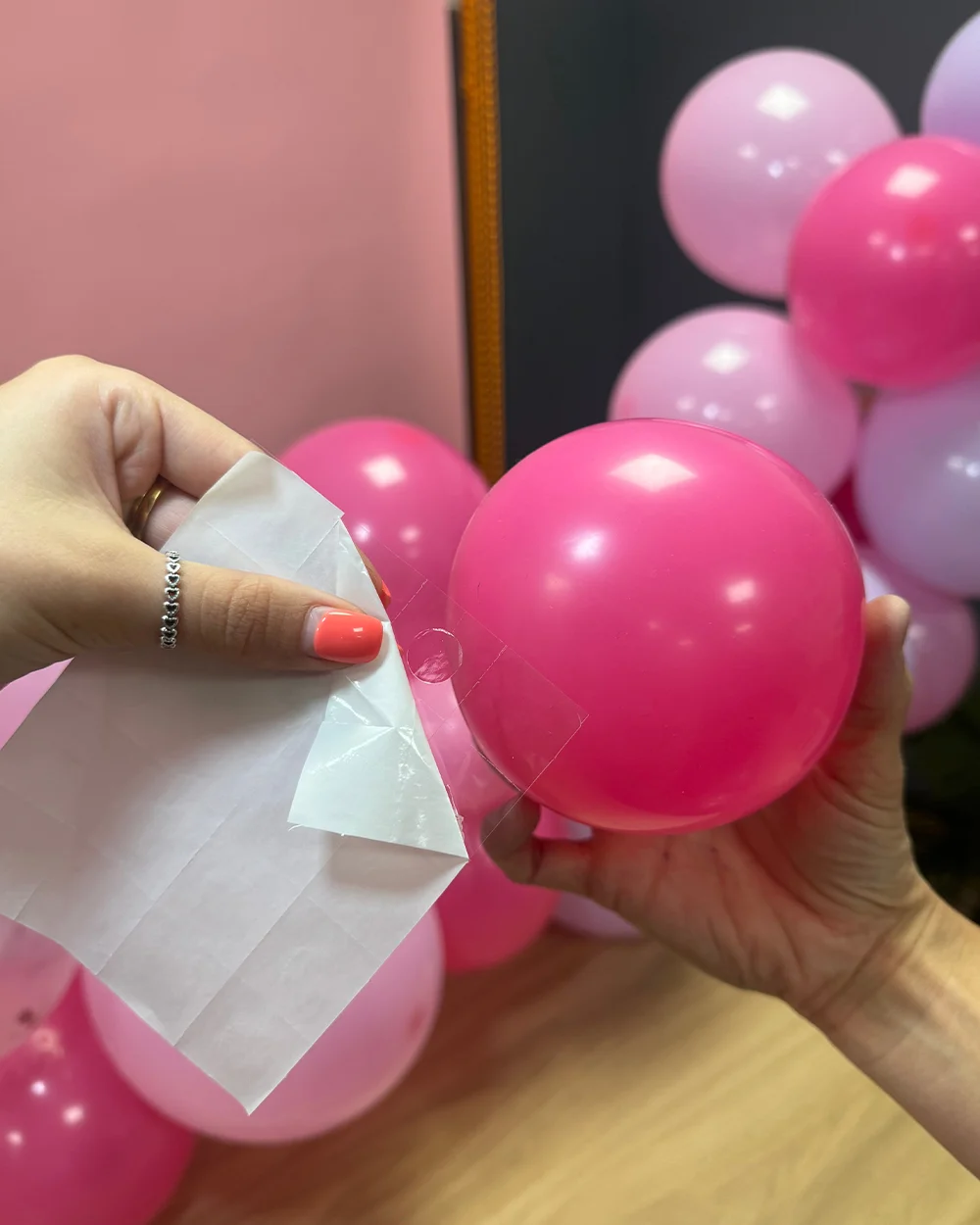 A close-up balloon arch tutorial step showing a small pink balloon being attached to a larger balloon arrangement using a clear adhesive glue dot. A hand peels back the adhesive backing while positioning the balloon against a cluster of pink, pale pink and lilac balloons, demonstrating how to fill gaps and add shape to a finished balloon arch. A close-up balloon arch tutorial step showing a small pink balloon being attached to a larger balloon arrangement using a clear adhesive glue dot. A hand peels back the adhesive backing while positioning the balloon against a cluster of pink, pale pink and lilac balloons, demonstrating how to fill gaps and add shape to a finished balloon arch.