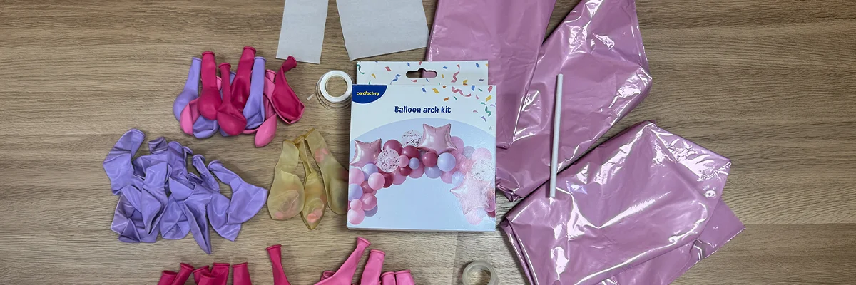 A balloon arch kit contents laid out on a wooden surface, including pink, lilac and gold balloons in assorted sizes, pink star-shaped foil balloons, adhesive tabs, clear tape, balloon arch strip, ribbon and instruction materials, arranged around the kit packaging showing the completed balloon arch design. A balloon arch kit contents laid out on a wooden surface, including pink, lilac and gold balloons in assorted sizes, pink star-shaped foil balloons, adhesive tabs, clear tape, balloon arch strip, ribbon and instruction materials, arranged around the kit packaging showing the completed balloon arch design.