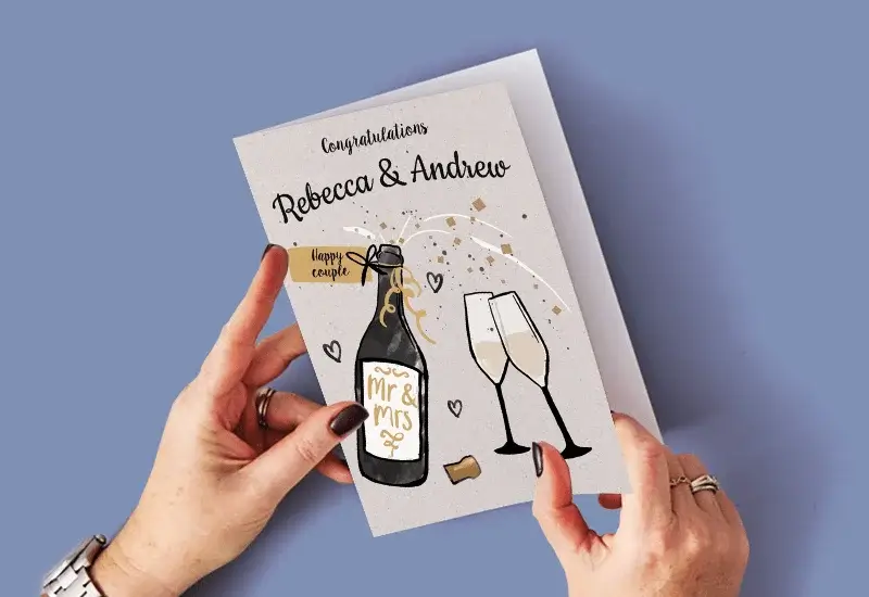 A person holding a wedding card with a champagne bottle labeled Mr and Mrs, two glasses, hearts, confetti, and the text Congratulations Rebecca and Andrew.