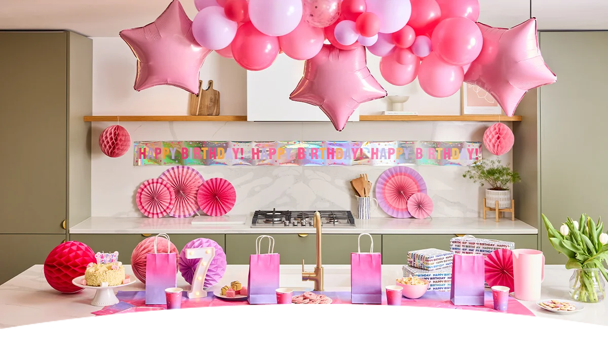 A pink-themed birthday party display set in a modern kitchen, featuring a large balloon garland of pink balloons and star-shaped foil balloons suspended above a table. A holographic ‘Happy Birthday’ banner and pink paper fan decorations adorn the background, while the table is styled with pink and purple ombré party bags, matching cups and plates, wrapped presents, snacks, a birthday cake on a stand, and a light-up number 7 decoration, creating a coordinated celebration setup. A pink-themed birthday party display set in a modern kitchen, featuring a large balloon garland of pink balloons and star-shaped foil balloons suspended above a table. A holographic ‘Happy Birthday’ banner and pink paper fan decorations adorn the background, while the table is styled with pink and purple ombré party bags, matching cups and plates, wrapped presents, snacks, a birthday cake on a stand, and a light-up number 7 decoration, creating a coordinated celebration setup.