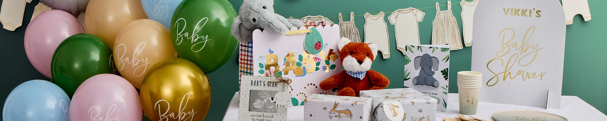 A baby shower display featuring pastel balloons in green, gold, blue and pink, personalised tableware, wrapped gifts and baby-themed keepsakes arranged on a table. Decorative baby clothes bunting hangs above, alongside a sign reading ‘Vikki’s Baby Shower’ and balloons printed with the word ‘Baby.’