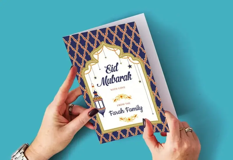 A person holding an ornate Eid Mubarak greeting card featuring gold and navy patterns, a lantern illustration, and decorative text, set against a purple background.