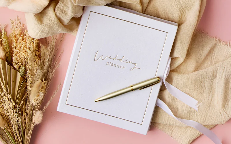 A white ‘Wedding Planner’ book with gold lettering and a matching gold pen resting on top, styled with dried flowers and beige fabric on a pink background. A white ‘Wedding Planner’ book with gold lettering and a matching gold pen resting on top, styled with dried flowers and beige fabric on a pink background.