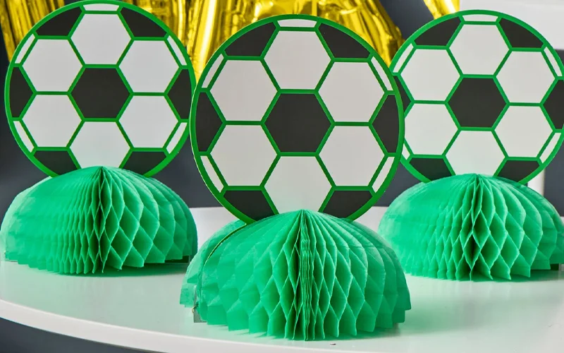 Three football‑themed honeycomb table centrepieces featuring green and black football designs mounted on green paper honeycomb bases, displayed in front of gold foil party decorations. Three football‑themed honeycomb table centrepieces featuring green and black football designs mounted on green paper honeycomb bases, displayed in front of gold foil party decorations.
