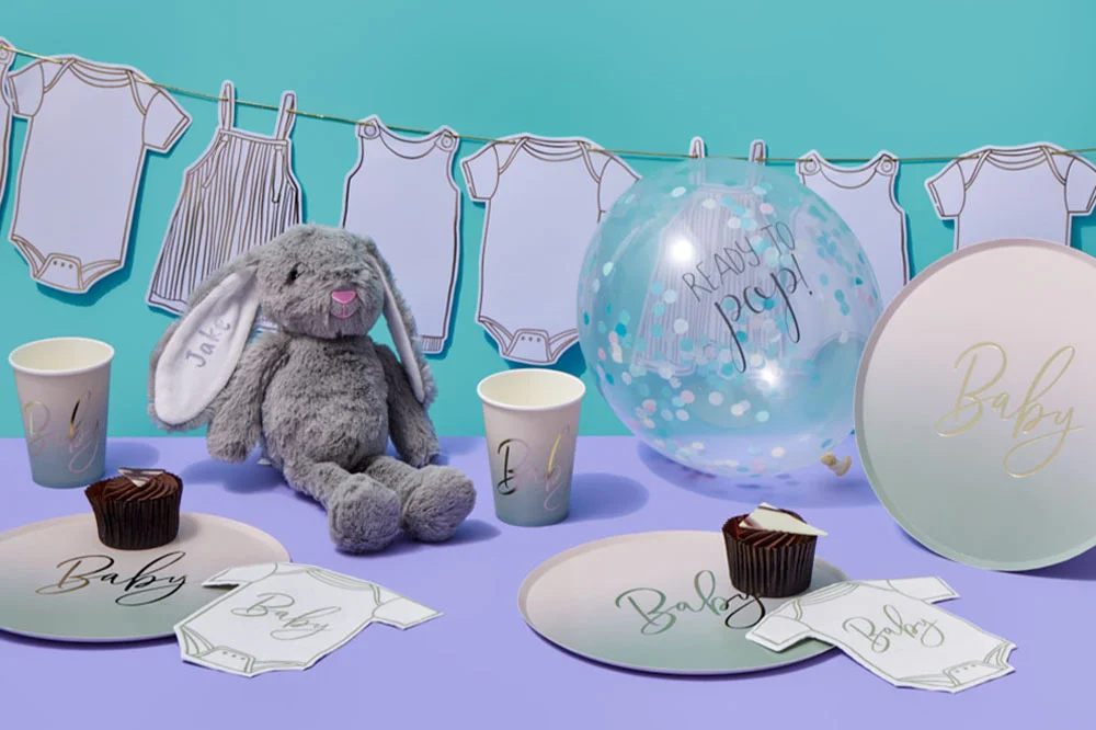 A personalised baby shower display with a grey bunny soft toy, a clear ‘Ready to Pop!’ confetti balloon, ombré baby-themed plates and cups, and baby bodysuit-shaped napkins arranged on a lilac table surface. Baby-clothes bunting hangs in the background, highlighting the coordinated personalised baby shower theme. A personalised baby shower display with a grey bunny soft toy, a clear ‘Ready to Pop!’ confetti balloon, ombré baby-themed plates and cups, and baby bodysuit-shaped napkins arranged on a lilac table surface. Baby-clothes bunting hangs in the background, highlighting the coordinated personalised baby shower theme.