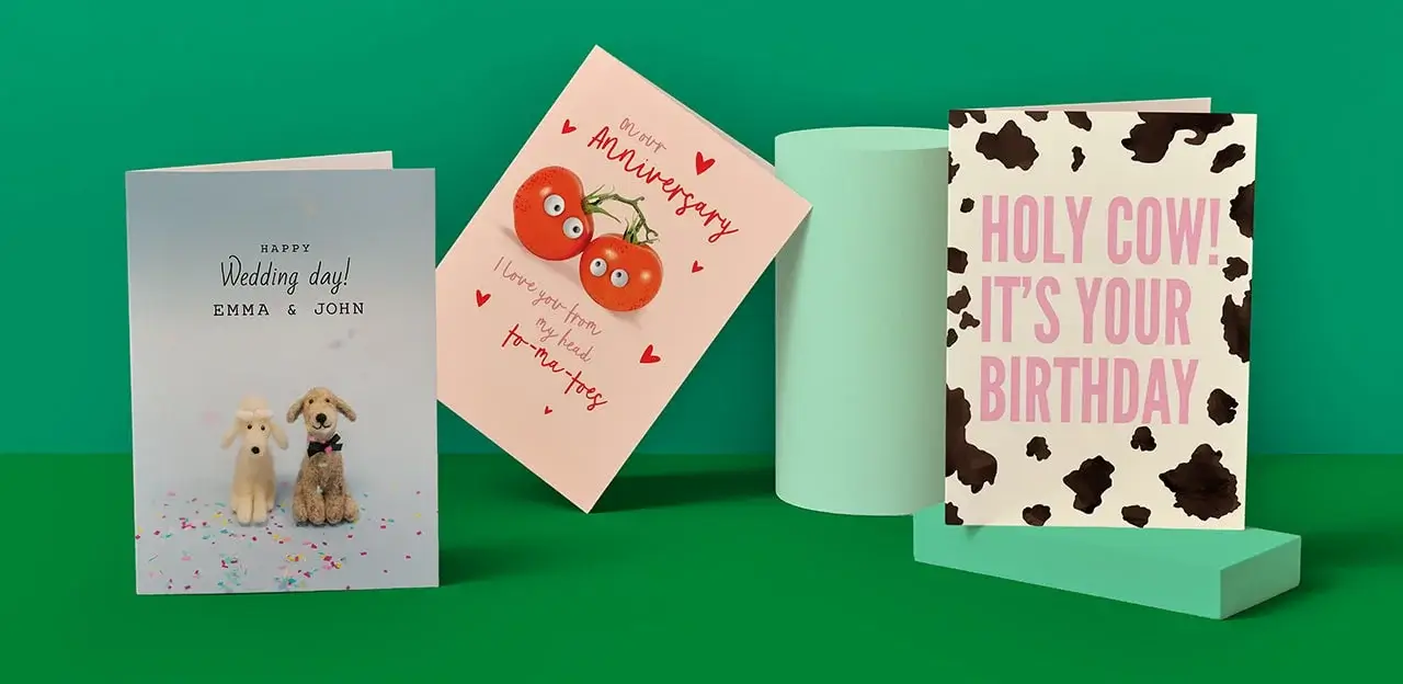 Three greeting cards on a green background: one with dogs saying 'Happy Wedding day! EMMA & JOHN', one with tomatoes saying 'On our Anniversary I love you from my head to-ma-toes', and one with a cow print saying 'HOLY COW! IT'S YOUR BIRTHDAY'
