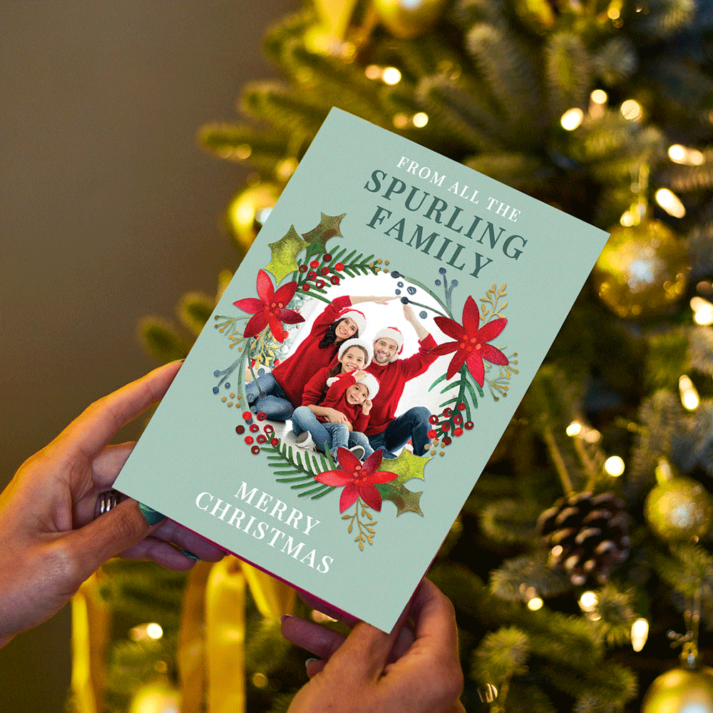 Christmas card held in front of a decorated tree, featuring a family photo framed by a festive wreath with red poinsettias and holly, with the text From all the Spurling Family at the top and Merry Christmas at the bottom.