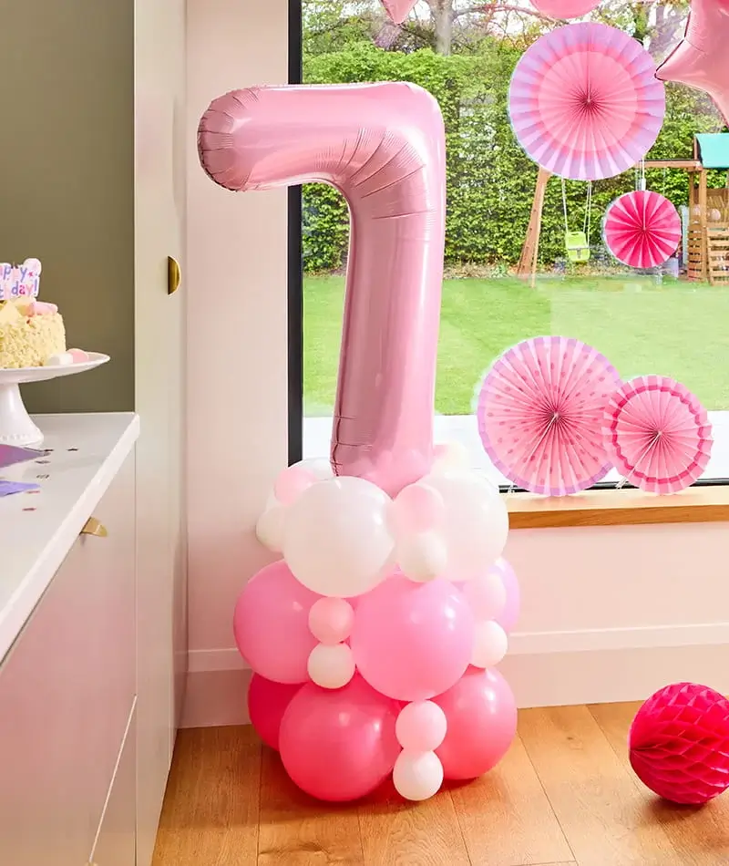 Pink number 7 balloon display with a balloon base, pink paper fan decorations, and birthday party décor beside a window. Pink number 7 balloon display with a balloon base, pink paper fan decorations, and birthday party décor beside a window.