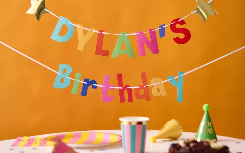 A colourful personalised birthday bunting banner displaying the name ‘Dylan’ and the word ‘Birthday’ in multicoloured letters, hung against a bright orange background above party tableware and decorations.