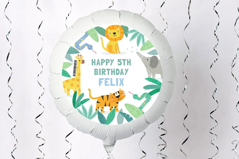 A round personalised foil balloon decorated with jungle animals including a lion, giraffe, tiger, elephant and snake, featuring the message ‘Happy 5th Birthday Felix’. The balloon is displayed against a white background with hanging silver streamers.