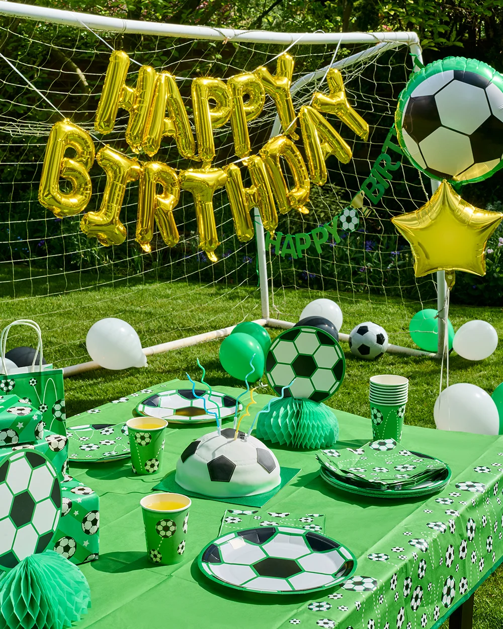A football-themed birthday party table set up outdoors in front of a goal net, decorated with green football-print tableware including plates, cups, napkins and a tablecloth. The display features a football-shaped birthday cake, wrapped presents, football honeycomb centrepieces, balloons, and a large gold ‘Happy Birthday’ balloon banner with football-themed balloon decorations. A football-themed birthday party table set up outdoors in front of a goal net, decorated with green football-print tableware including plates, cups, napkins and a tablecloth. The display features a football-shaped birthday cake, wrapped presents, football honeycomb centrepieces, balloons, and a large gold ‘Happy Birthday’ balloon banner with football-themed balloon decorations.