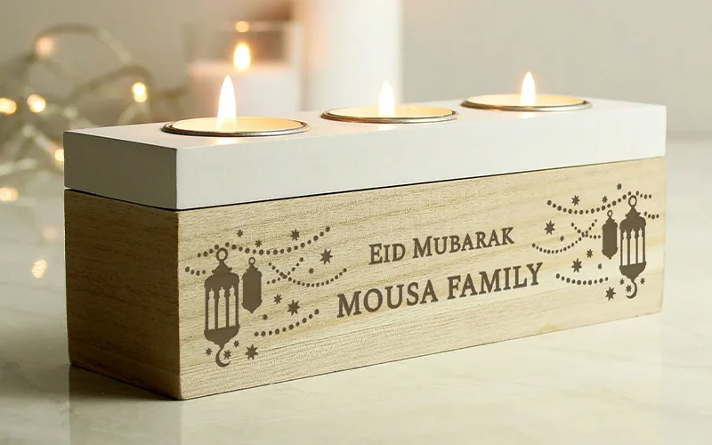A wooden tealight holder with three lit candles on top, engraved with lanterns, stars, and the text ‘Eid Mubarak Mousa Family.
