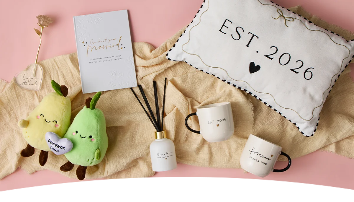 A flat‑lay of wedding‑themed gifts arranged on a soft beige fabric over a pink background, including an ‘Our First Year Married’ journal, pear plush toys holding a ‘Perfect Pear’ heart, a reed diffuser, matching mugs printed ‘EST. 2026,’ and a decorative cushion with ‘EST. 2026’ and a heart design. A flat‑lay of wedding‑themed gifts arranged on a soft beige fabric over a pink background, including an ‘Our First Year Married’ journal, pear plush toys holding a ‘Perfect Pear’ heart, a reed diffuser, matching mugs printed ‘EST. 2026,’ and a decorative cushion with ‘EST. 2026’ and a heart design.