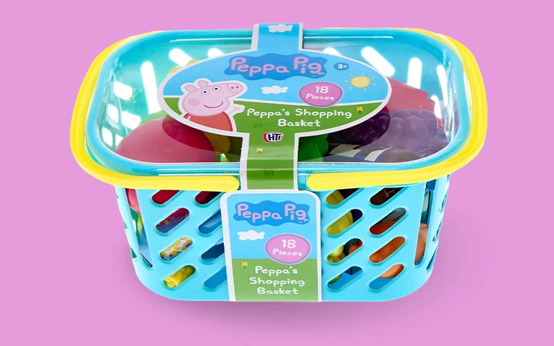 A Peppa Pig pretend shopping basket toy with a blue plastic basket and assorted play food items inside, shown in branded packaging on a pink background.