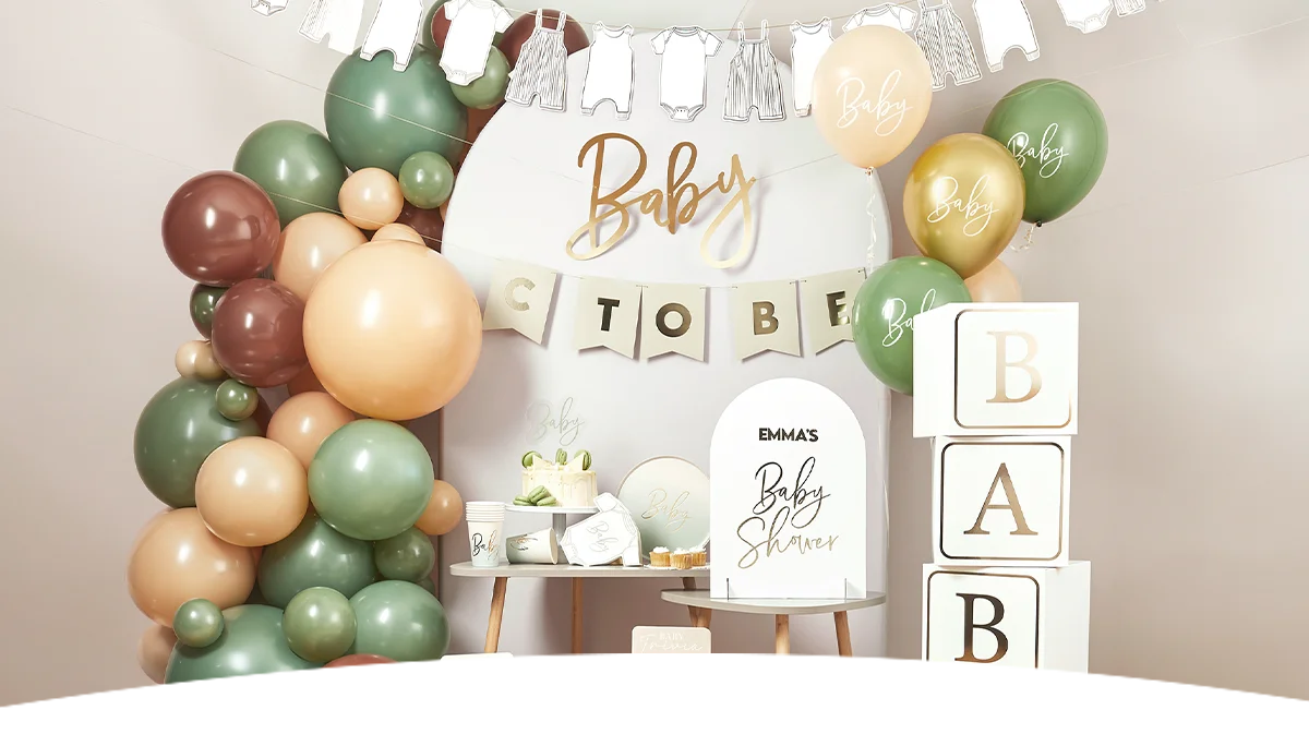 A baby shower celebration display with a sage green, cream and mocha balloon garland, a gold ‘Baby’ backdrop, a ‘Baby to Be’ bunting banner, personalised ‘Emma’s Baby Shower’ signage, coordinating tableware, and stacked decorative blocks spelling ‘BABY.’ White baby clothes bunting hangs above the display, creating a stylish baby shower setup.