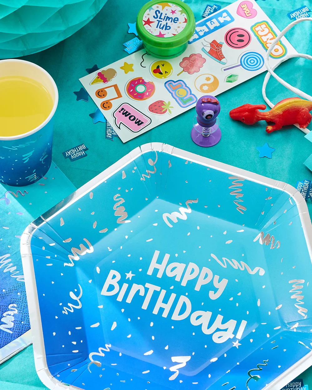 A blue hexagonal party plate featuring white confetti-style details and bold text reading ‘Happy Birthday!’ in the centre. The plate is displayed on a turquoise party table alongside a matching blue cup, birthday-themed confetti, a sticker sheet, a tub of slime, and small party bag fillers. A blue hexagonal party plate featuring white confetti-style details and bold text reading ‘Happy Birthday!’ in the centre. The plate is displayed on a turquoise party table alongside a matching blue cup, birthday-themed confetti, a sticker sheet, a tub of slime, and small party bag fillers.