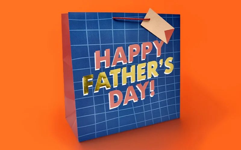 A Father’s Day gift bag with a blue grid pattern design, featuring bold multicoloured lettering reading ‘Happy Father’s Day!’ and a small tag attached to the handle, displayed against a bright orange background. A Father’s Day gift bag with a blue grid pattern design, featuring bold multicoloured lettering reading ‘Happy Father’s Day!’ and a small tag attached to the handle, displayed against a bright orange background.