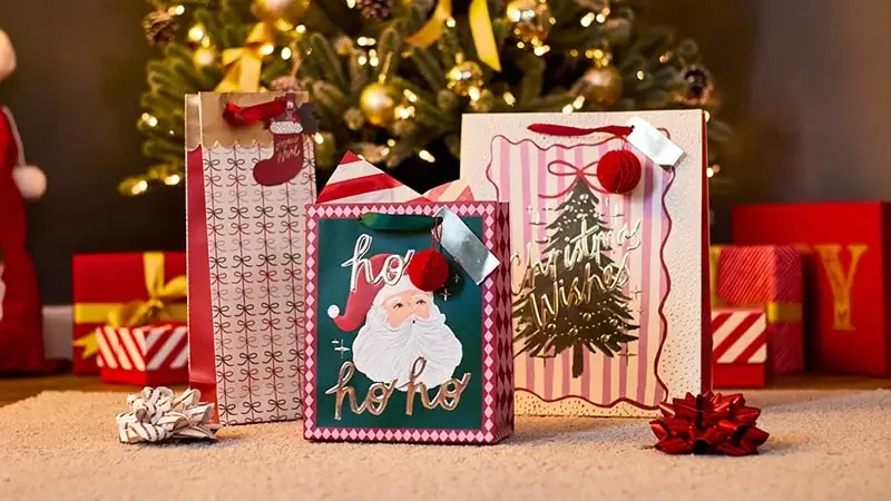 Christmas gift bags with festive designs, including Santa and a Christmas tree, placed on a carpet in front of a decorated tree with wrapped presents and bows