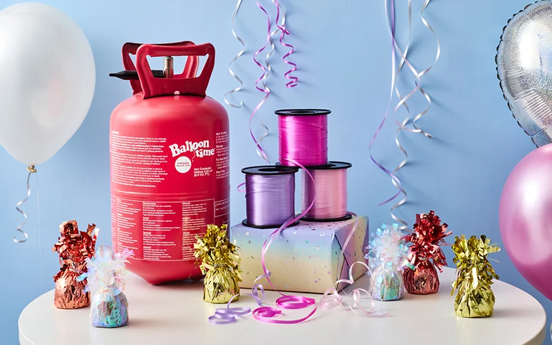 A balloon decorating display featuring a red helium tank, reels of curling ribbon in pink and purple shades, gift weights wrapped in metallic foil, and decorative balloon accessories arranged on a table against a pale blue background with hanging ribbons and balloons. A balloon decorating display featuring a red helium tank, reels of curling ribbon in pink and purple shades, gift weights wrapped in metallic foil, and decorative balloon accessories arranged on a table against a pale blue background with hanging ribbons and balloons.