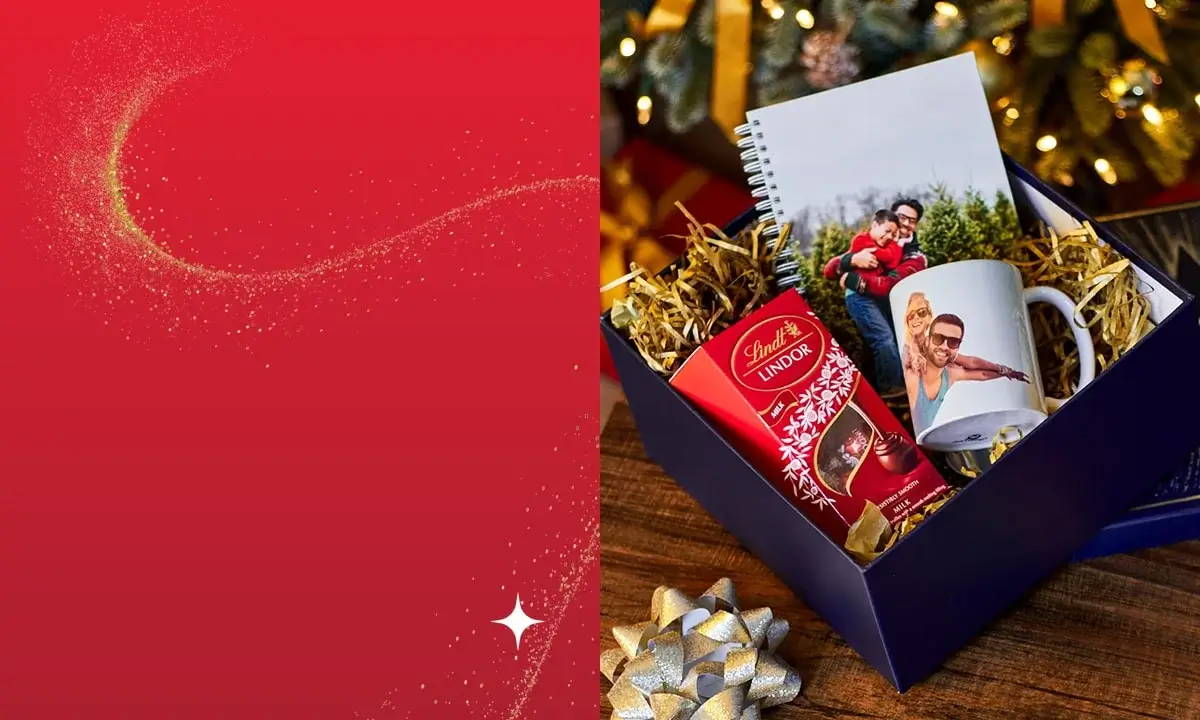 Gift box containing Lindt chocolates, a custom photo mug, and a spiral notebook, placed on a wooden surface with wrapped presents and a Christmas tree in the background.